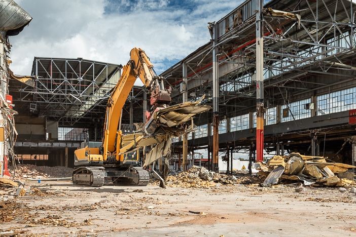 Yellow excavator demolishing an industrial building with large steel beams and rubble scattered around.